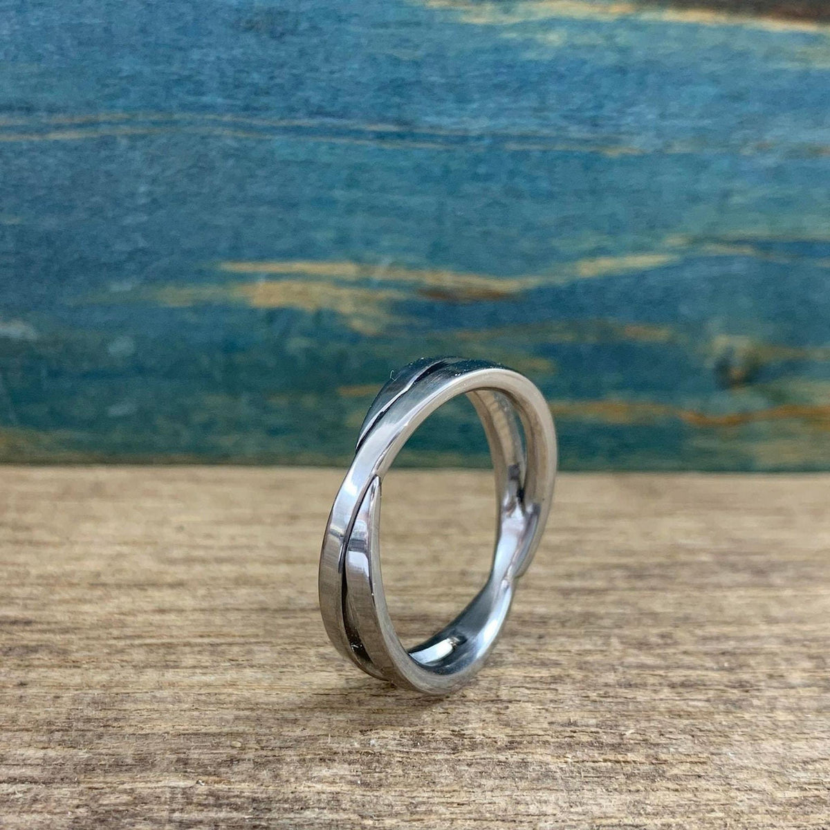 A shiny silver-colored crossover ring with two bands is shown from a low angle, resting on a light wooden surface.