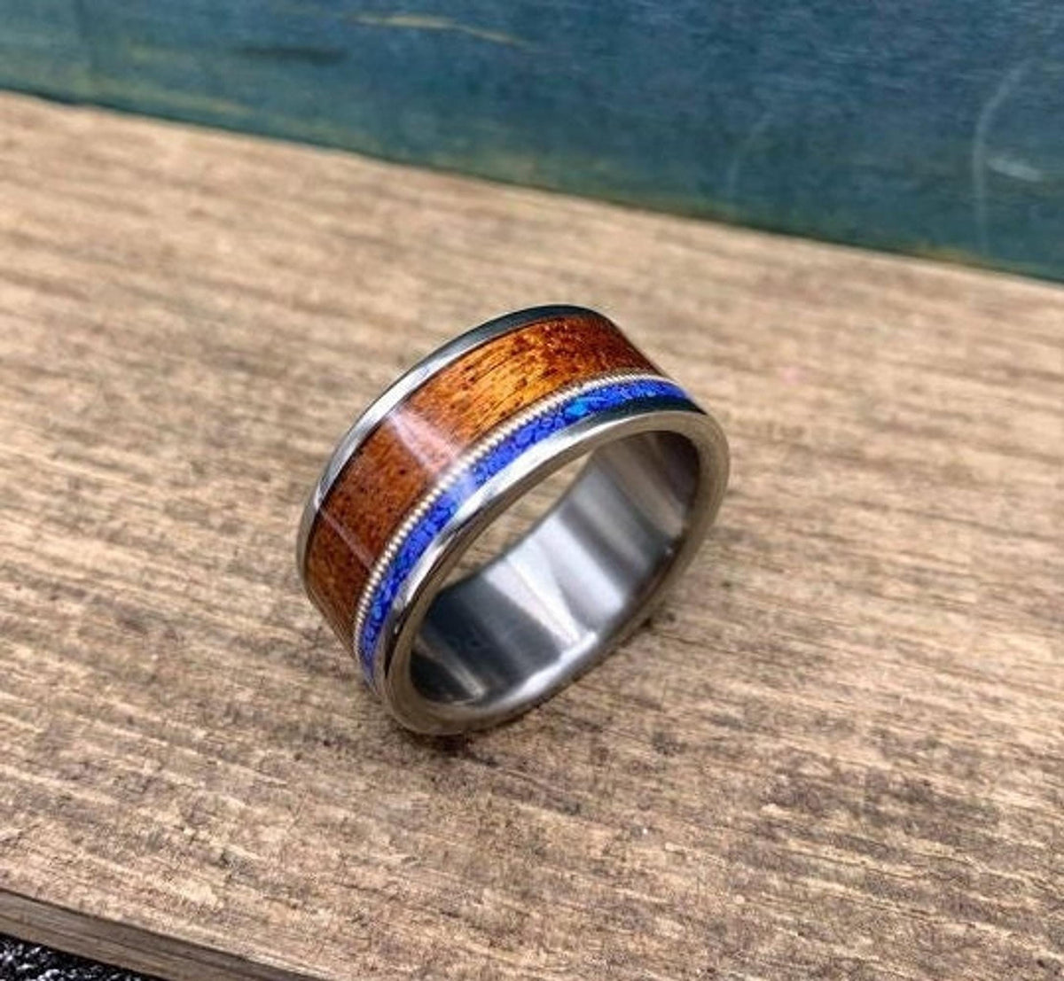 A silver-colored ring with a central dark wood inlay, a thin blue lapis lazuli inlay, and a guitar string inlay, on a light wood surface.