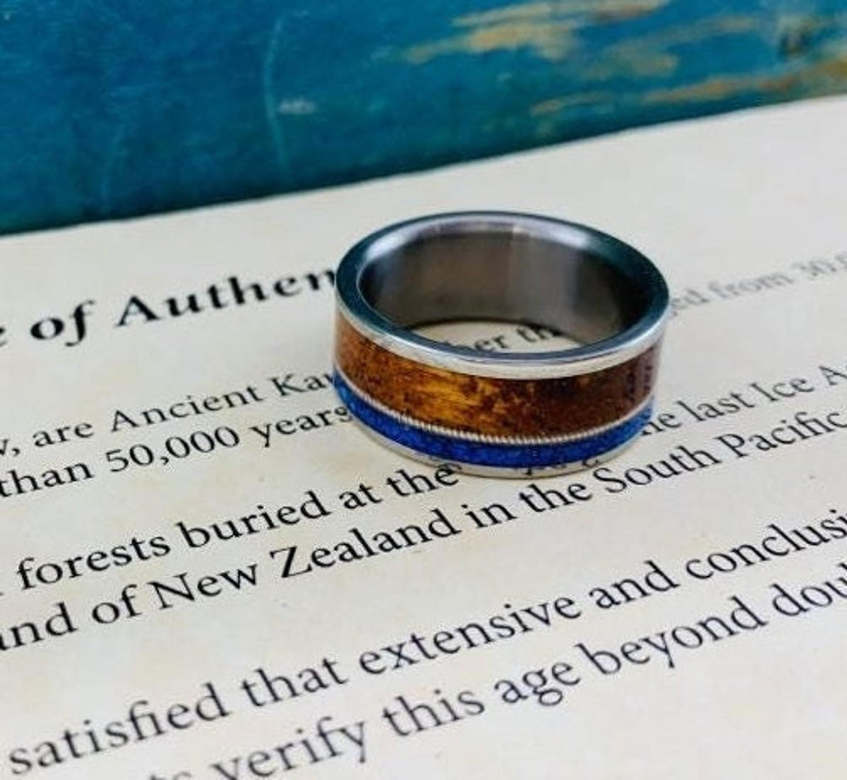 A silver-colored ring with a central dark wood inlay and a thin blue lapis lazuli inlay, resting on a document with text.