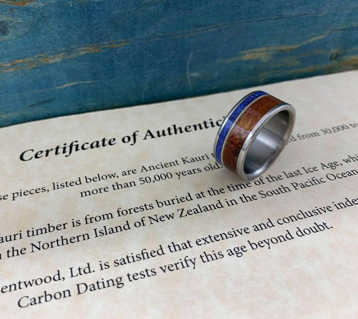 A silver-colored ring with a central dark wood inlay and a thin blue lapis lazuli inlay, resting on a document titled "Certificate of Authenticity."