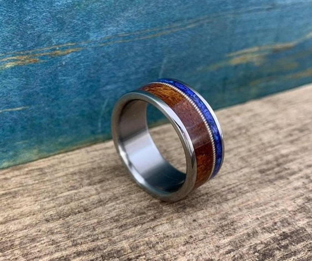 A silver-colored ring with a central dark wood inlay, a thin blue lapis lazuli inlay, and a guitar string inlay, on a light wood surface.