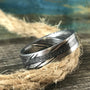 A silver-toned ring with a central textured band and patterned sides rests on a coil of rope on a wooden surface.