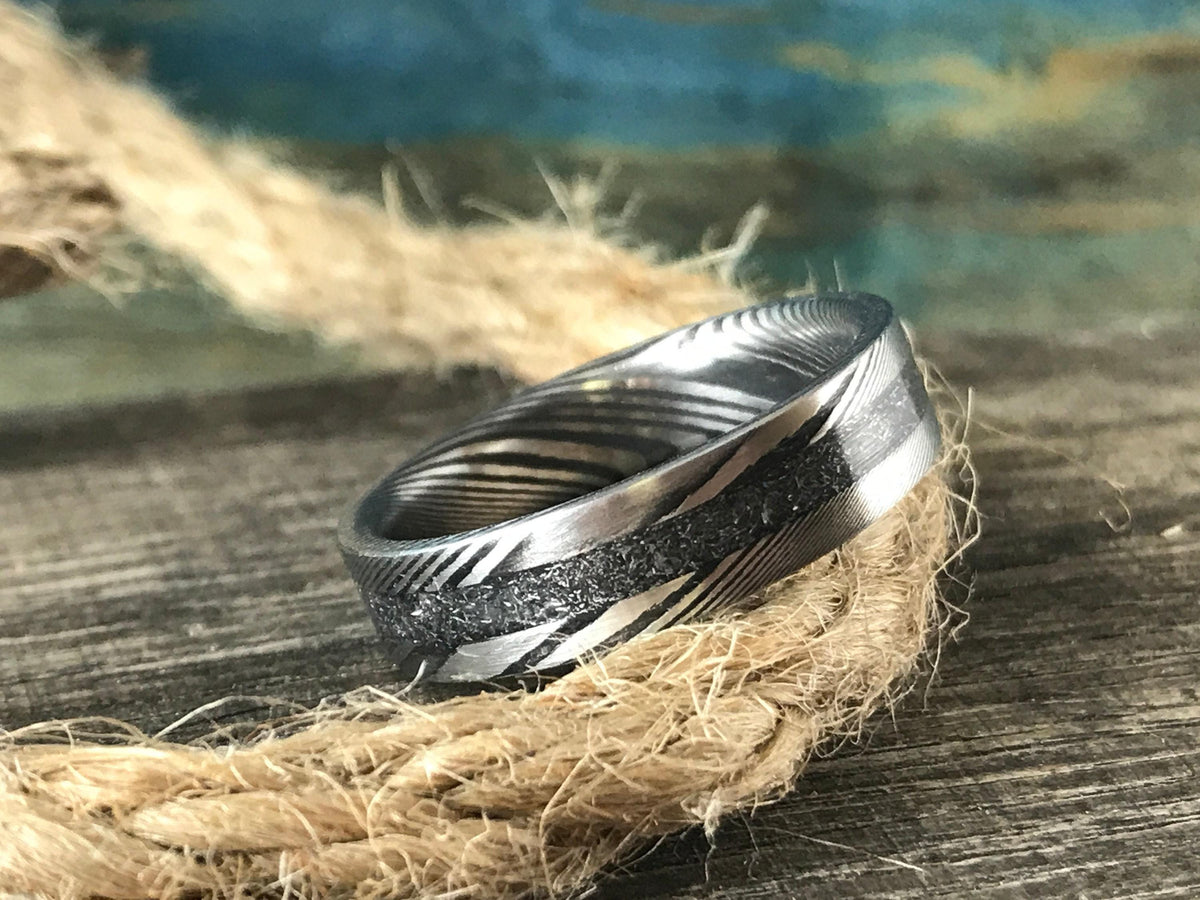 A silver-toned ring with a central textured band and patterned sides rests on a coil of rope on a wooden surface.