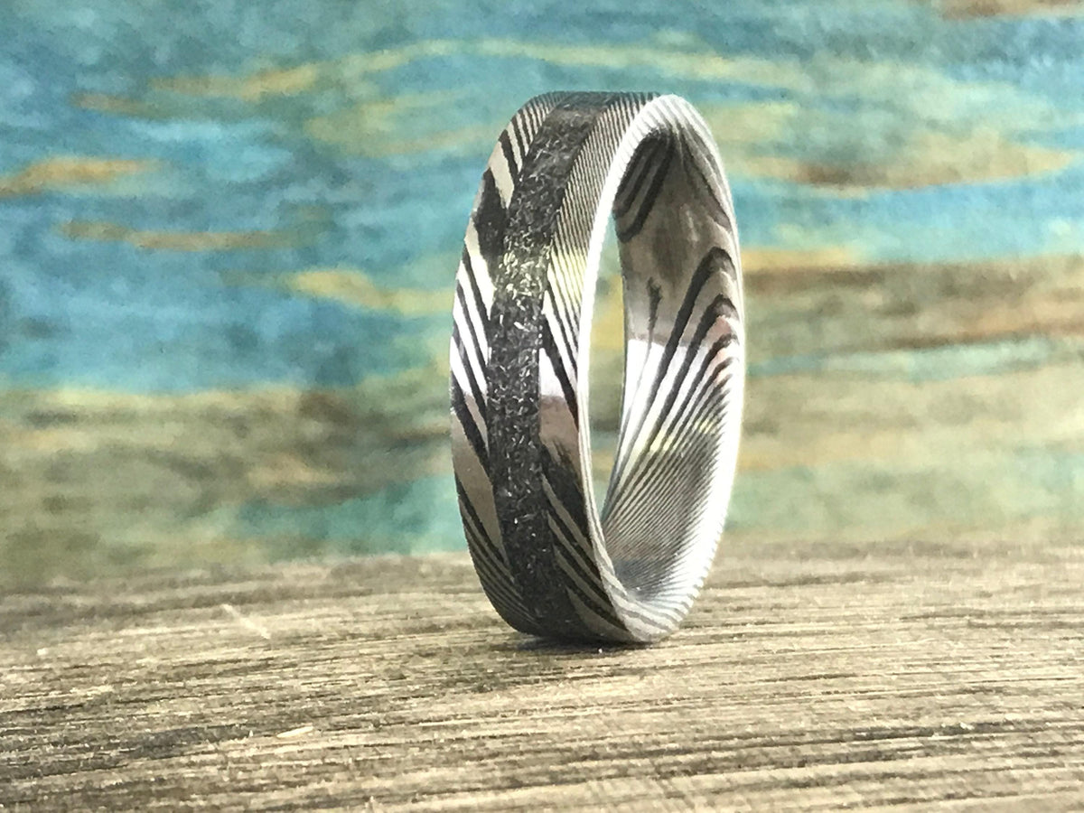 A silver-toned ring with a central textured band and patterned sides stands upright on a wooden surface, angled slightly.