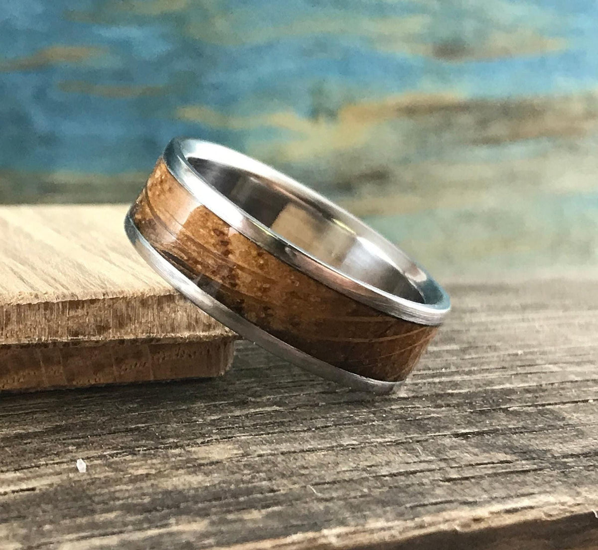 A silver-colored ring with a light brown wood inlay, showing horizontal grain, is angled on a light brown wooden block with a blue and green background.