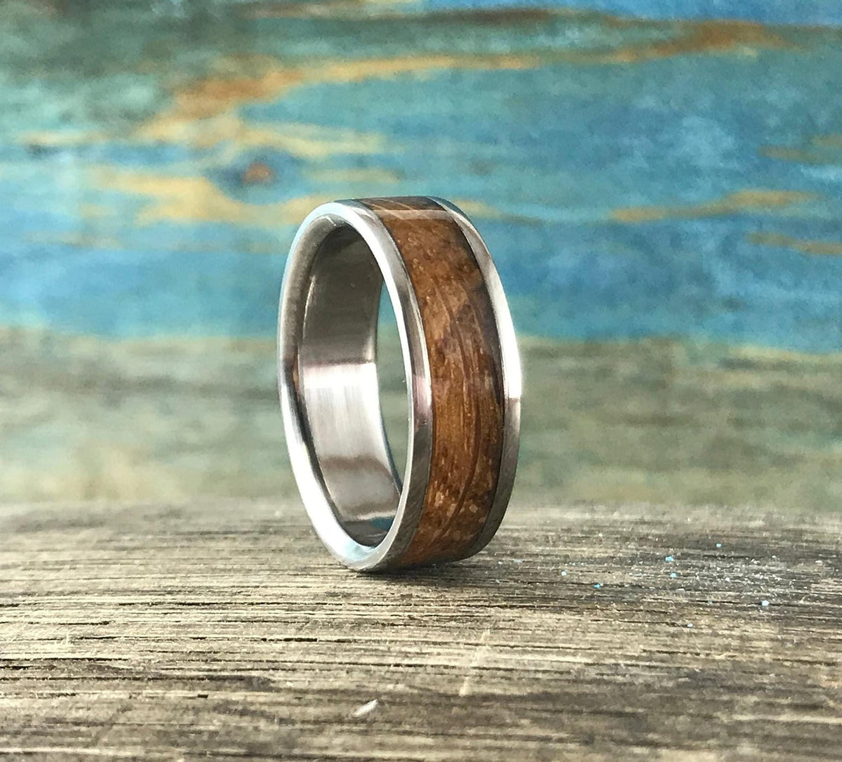 A silver-colored ring with a smooth, light brown wood inlay is centered on a light brown wooden surface with a blue and green background.