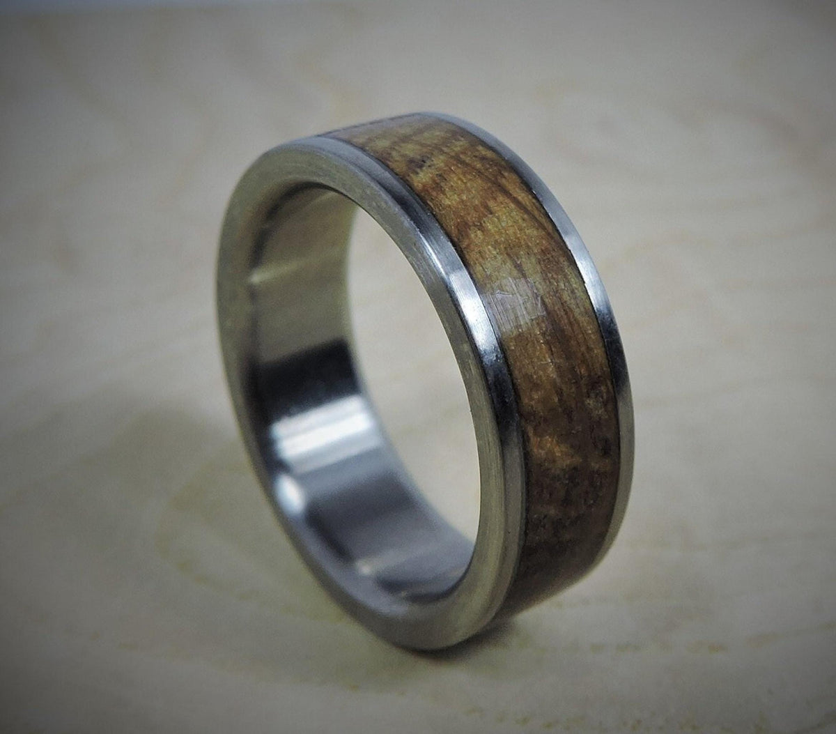 A silver-colored ring with a light brown wood inlay, showing subtle grain, is angled on a light wooden surface.