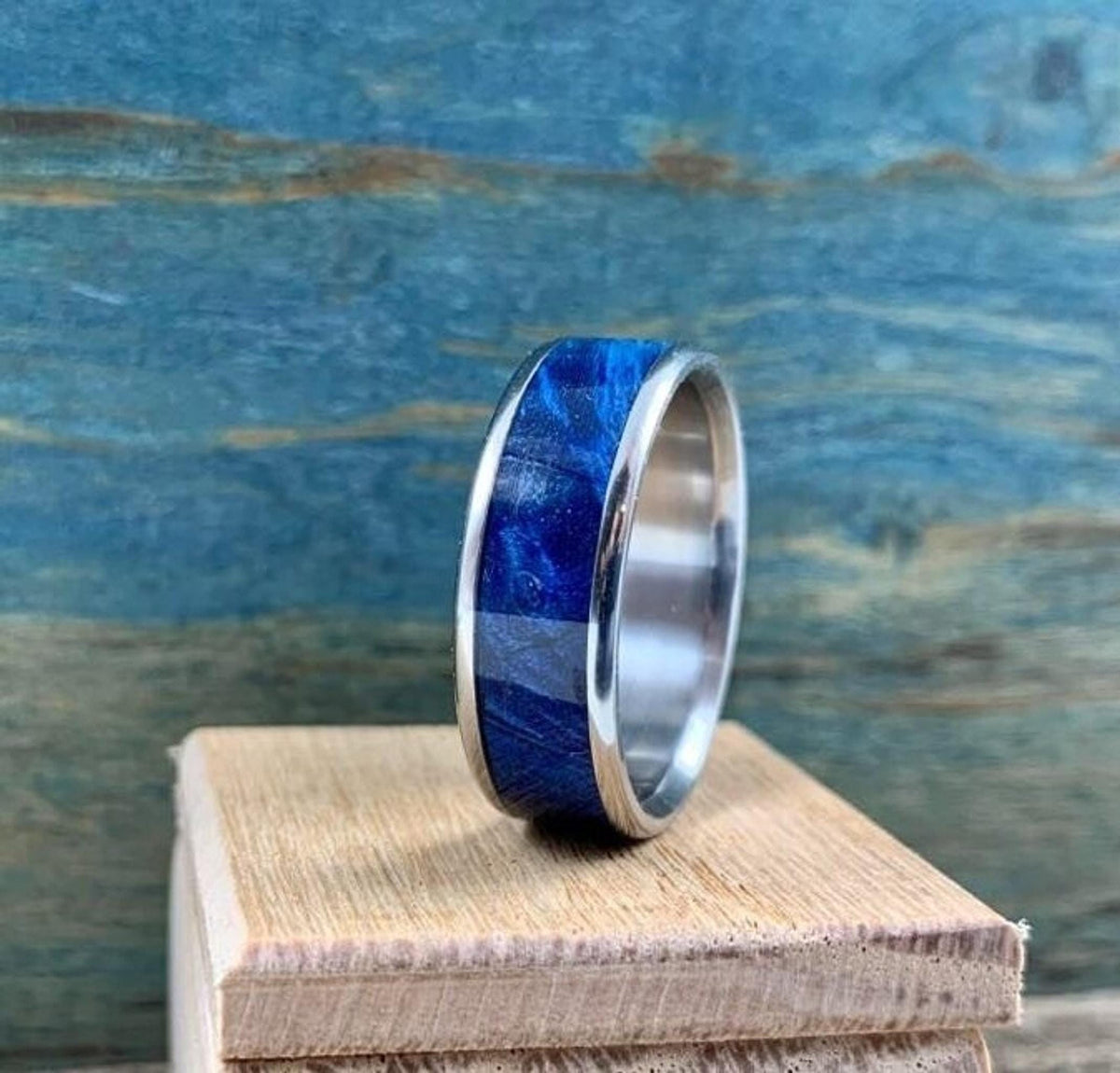 A silver-colored titanium ring with a blue box elder wood inlay stands upright on a light brown wooden block, with a blue wooden background.