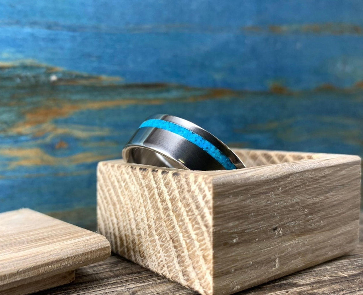 A silver-colored ring with a turquoise inlay, angled on a light wooden block with a blue background.