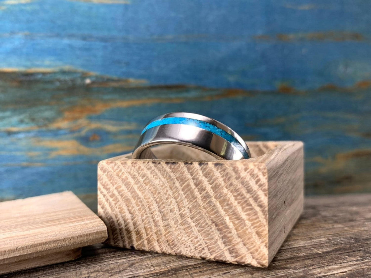 A silver-colored ring with a turquoise inlay, resting on a light wooden block with a blue background.