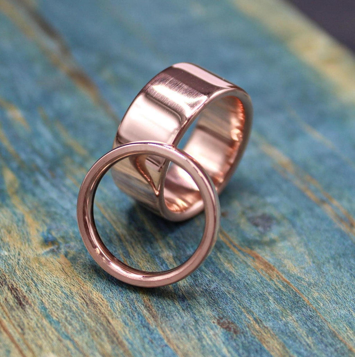 A narrow, shiny, rose gold-colored ring is positioned in front of a wider, matching ring on a blue-green distressed wooden surface.