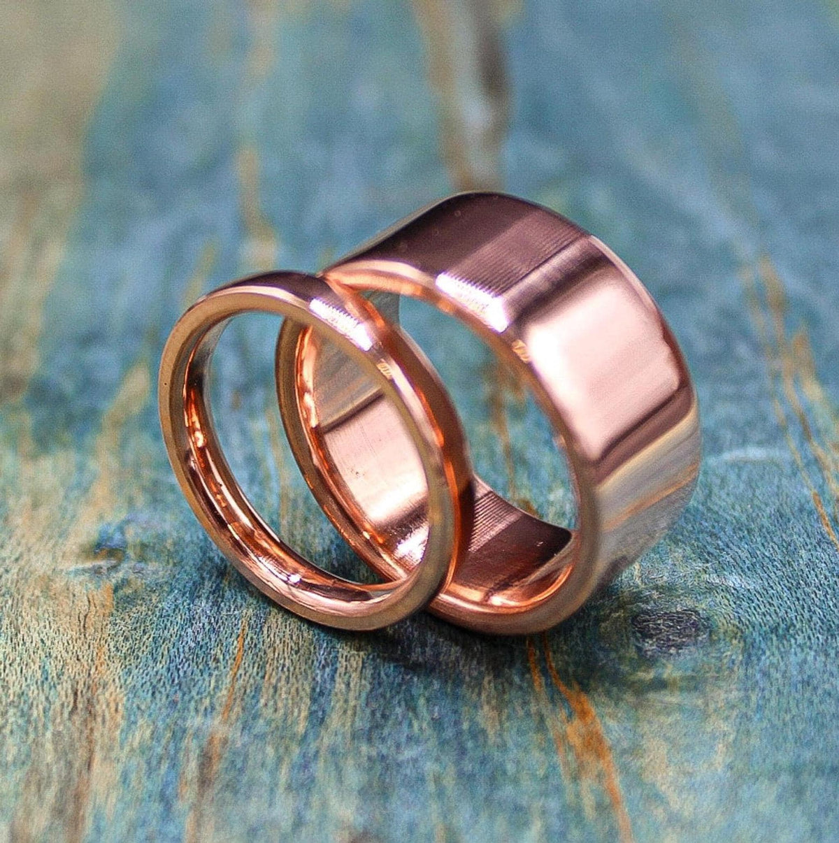 Two shiny, rose gold-colored rings, one thin and one wide, are stacked on a blue-green distressed wooden surface.