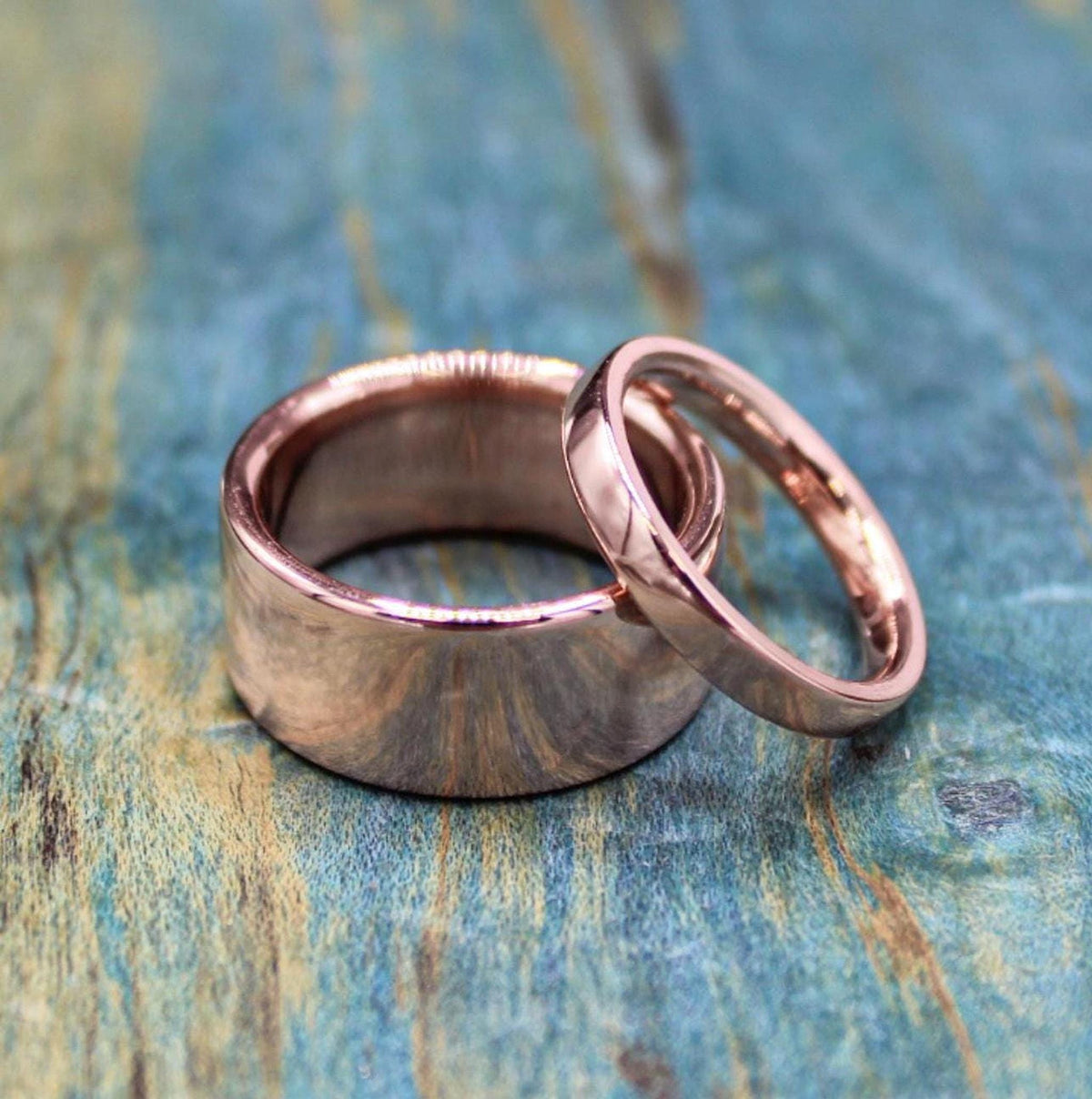A wide, shiny, rose gold-colored ring stands upright with a narrower, matching ring leaning against it on a blue-green distressed wooden surface.