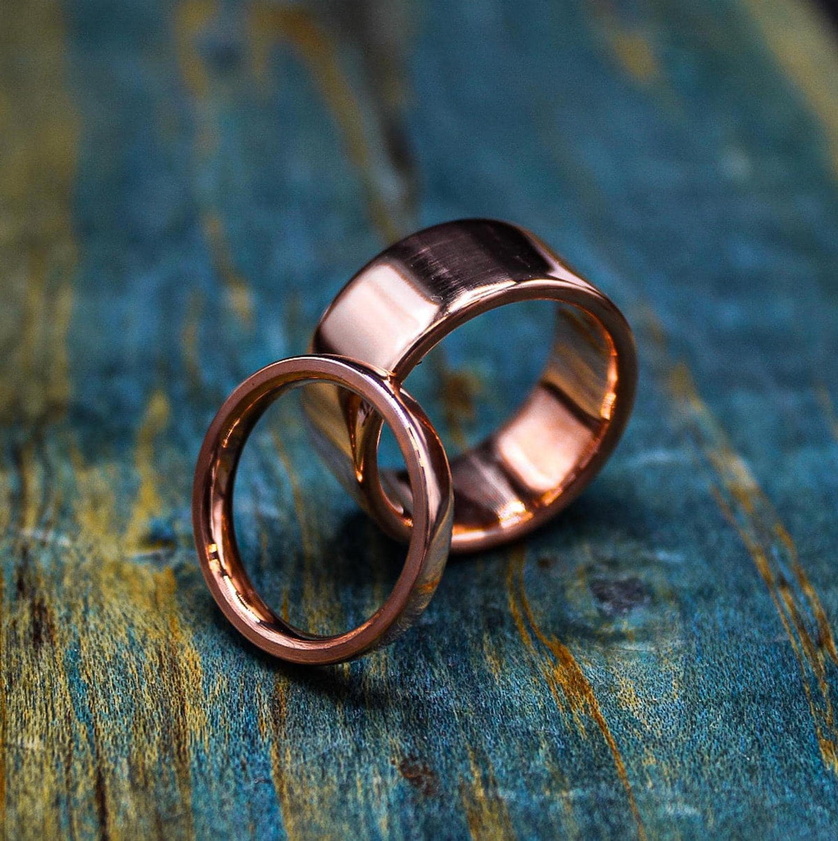 A thin, shiny, rose gold-colored ring is placed next to a wider, matching ring on a blue-green distressed wooden surface.
