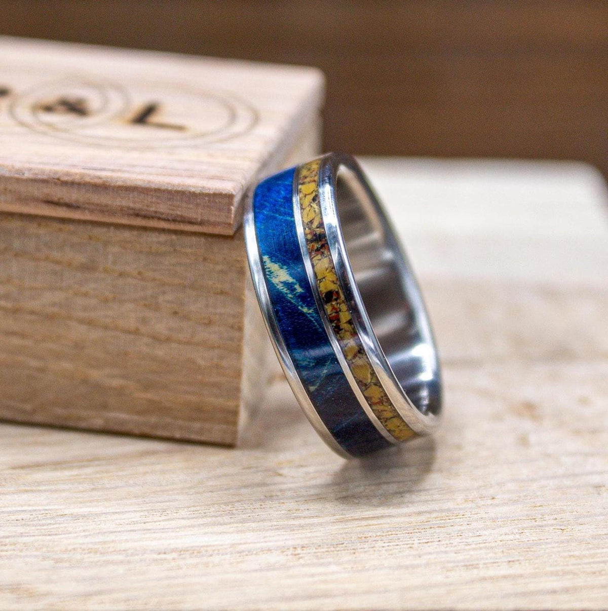 A silver-colored ring with a central band of yellow and blue material, flanked by blue material, rests on a light wooden surface next to a wooden box.