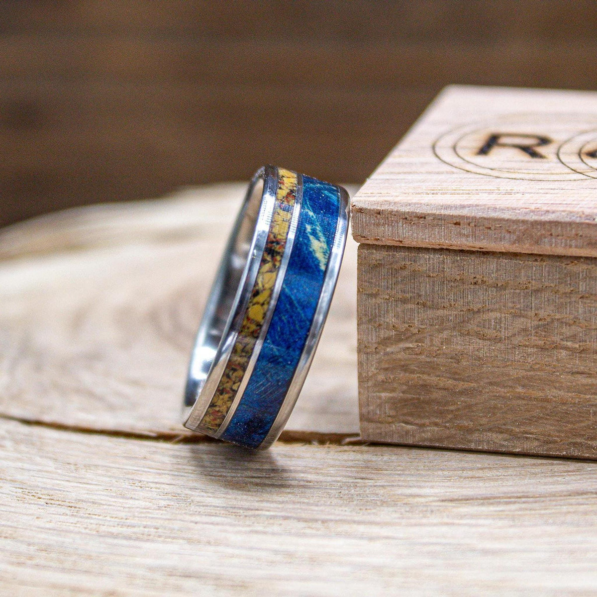 A silver-colored ring with a yellow and blue central band is angled on a light wooden surface, with a wooden box showing a circular logo in the background.