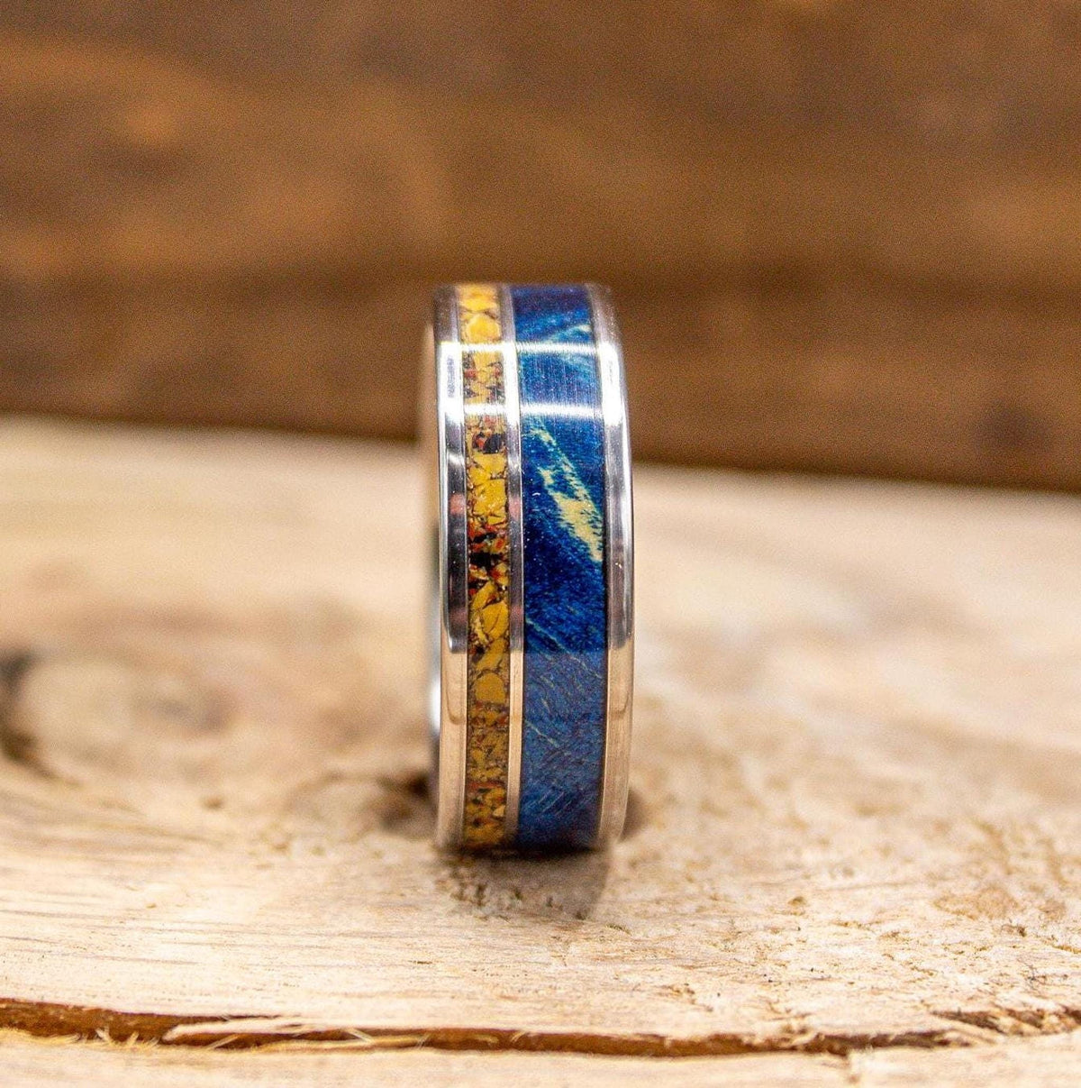 A silver-colored ring with a central yellow band and outer blue bands is shown from a straight-on perspective on a light wooden surface.