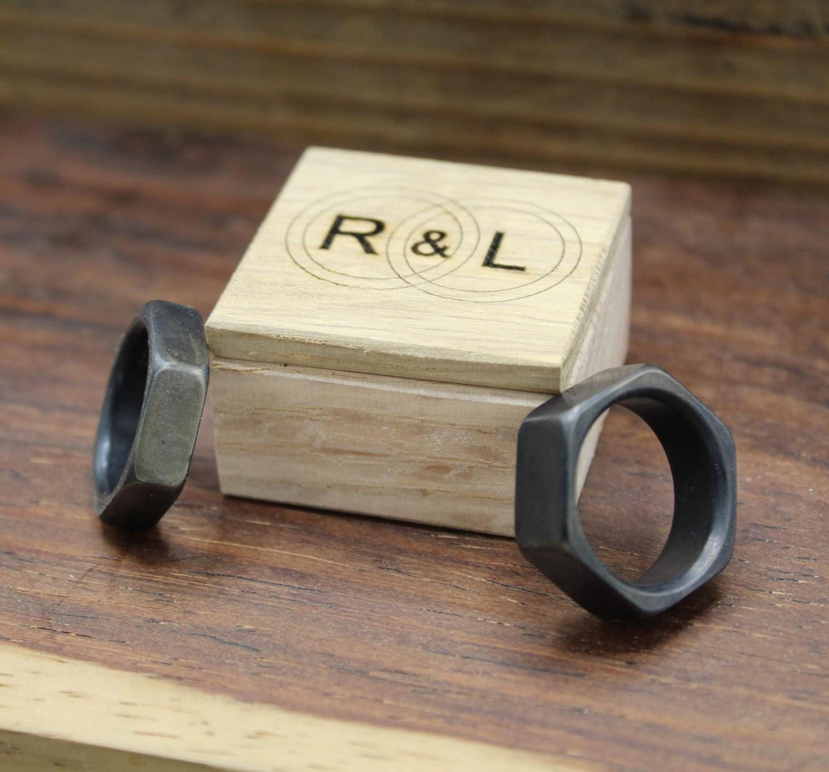 Two dark gray hexagonal rings flanking a light wooden box with "R & L" engraved on its lid, on a wooden surface.