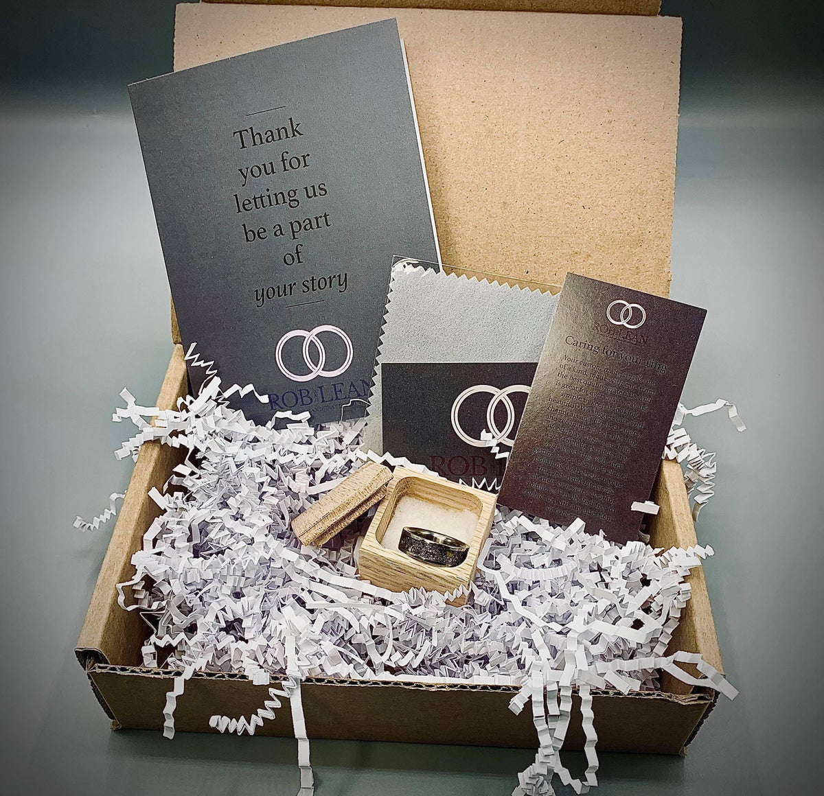 An open cardboard box contains white shredded paper, a small ring box, a gray card, and two black cards.