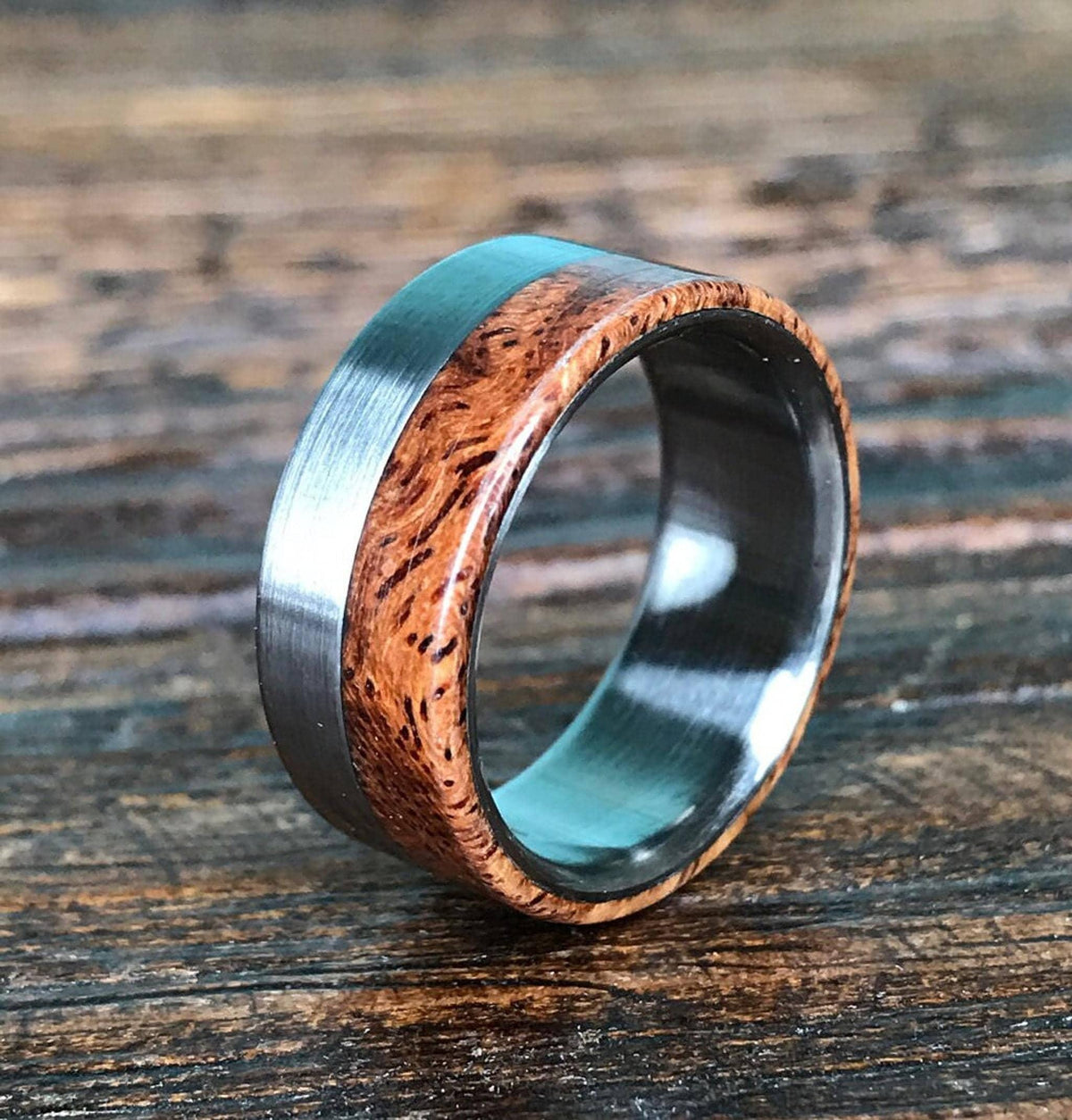 A ring with a brushed silver-colored metal band on the left, a reddish-brown wooden inlay with dark grain in the middle, and a polished silver-colored metal band on the right, on a dark wooden surface.