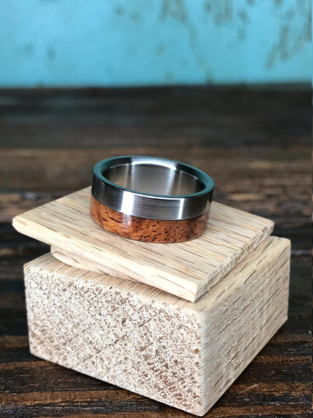 A ring with a polished silver-colored metal band and a reddish-brown wooden inlay with dark grain, resting on two light-colored wooden blocks, against a light blue wall.