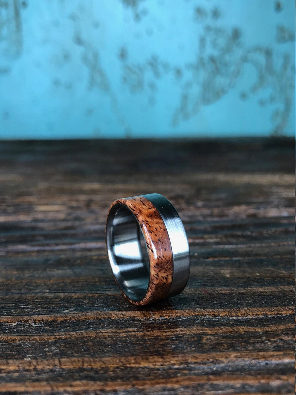 A ring with a brushed silver-colored metal band on the left, a reddish-brown wooden inlay with dark grain in the middle, and a polished silver-colored metal band on the right, on a dark wooden surface.