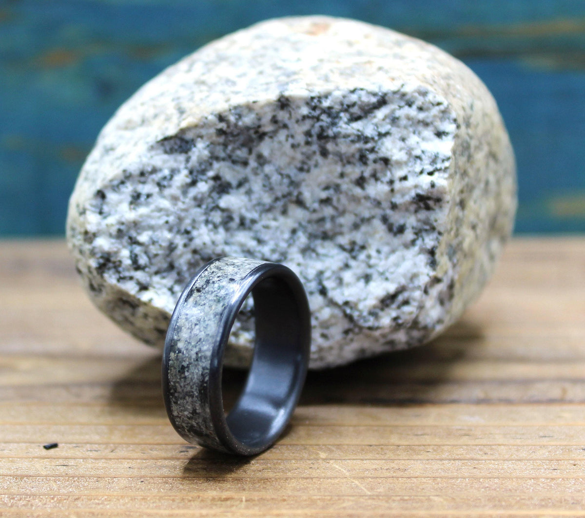 A black ring with a light gray, speckled granite inlay rests on a wooden surface, with a large, light gray granite rock in the background.