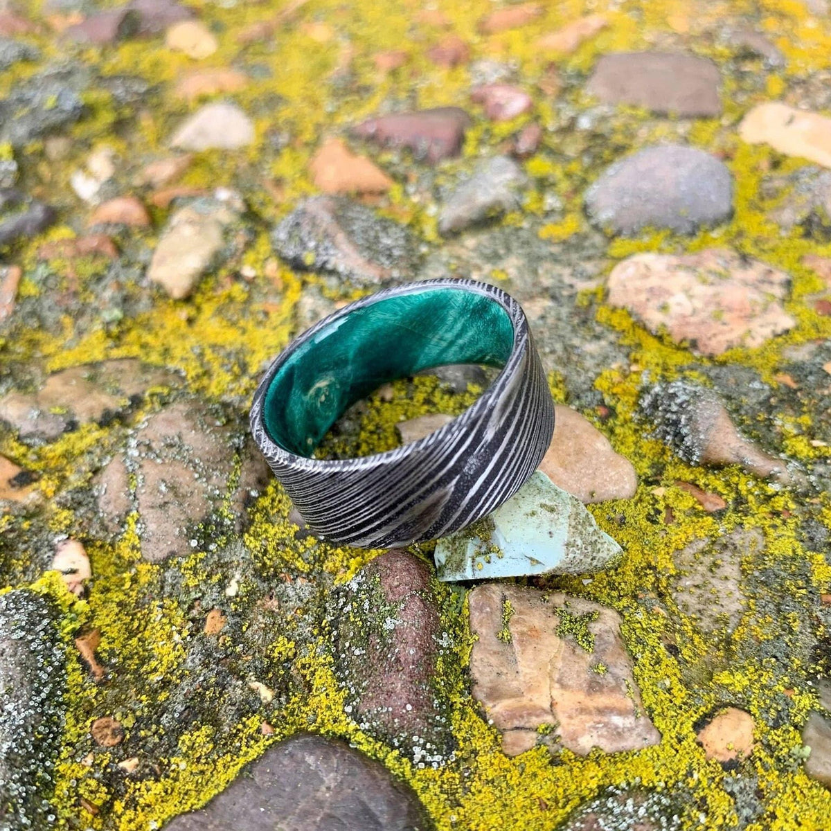 A wide, flat, patterned metal ring with a bright green interior is shown from a slightly different angle on mossy rocks, with a small blue-green rock next to it.