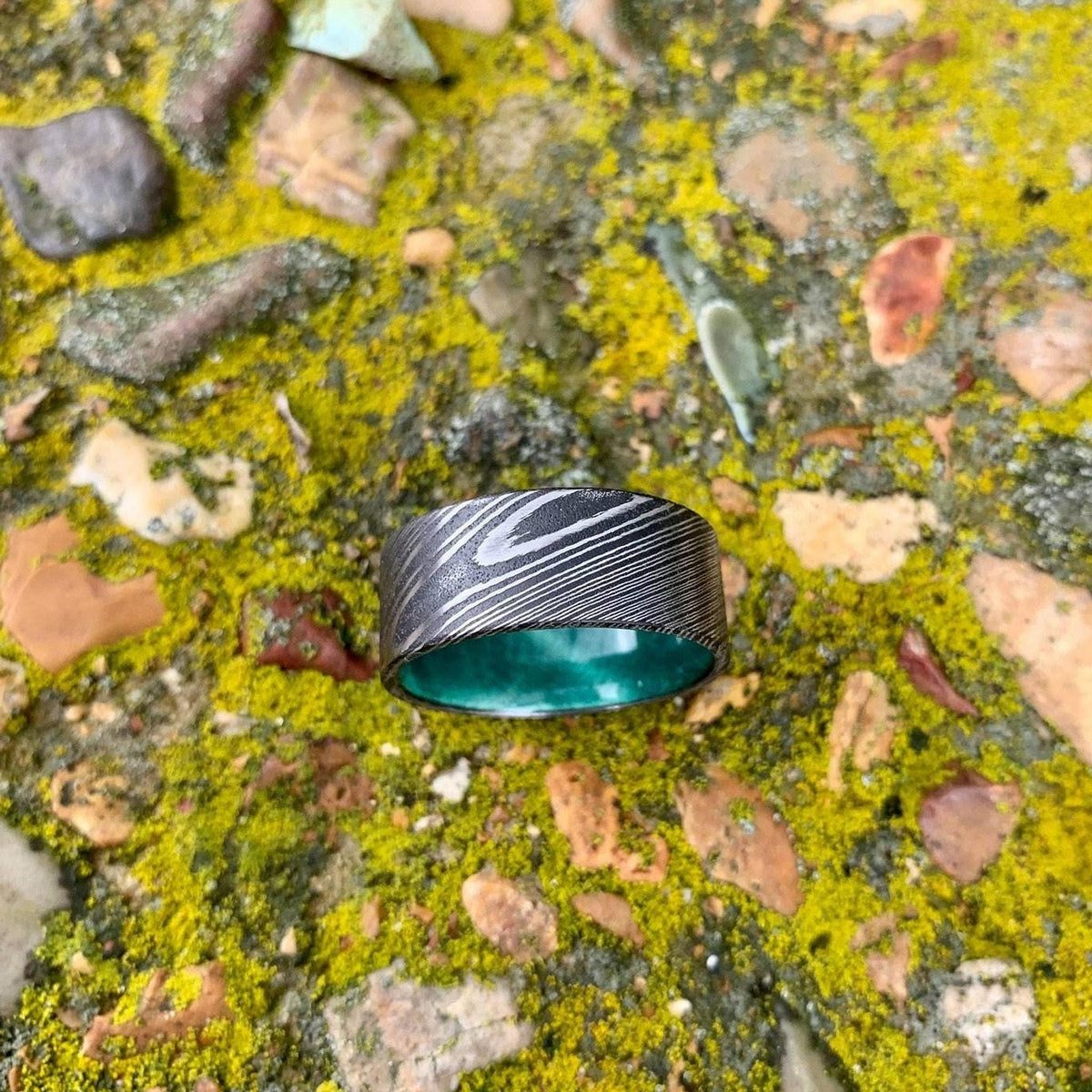 A wide, flat, patterned metal ring with a bright green interior is shown from above on mossy rocks.