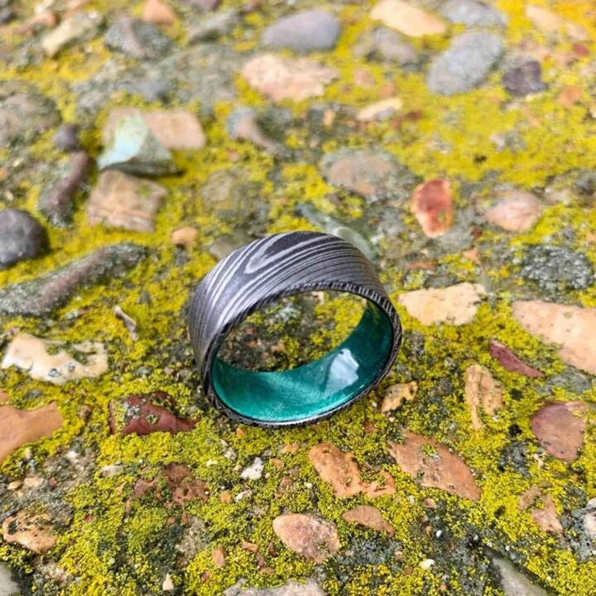 A wide, flat, patterned metal ring with a bright green interior is shown from above on mossy rocks, with more light on the ring.