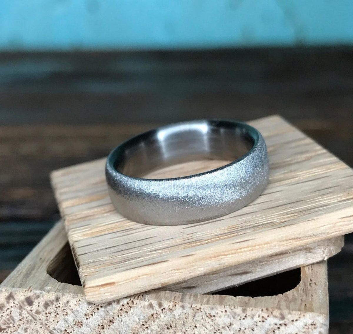 A silver-colored ring with a textured exterior and a smooth interior, displayed on light wooden blocks against a blue background.