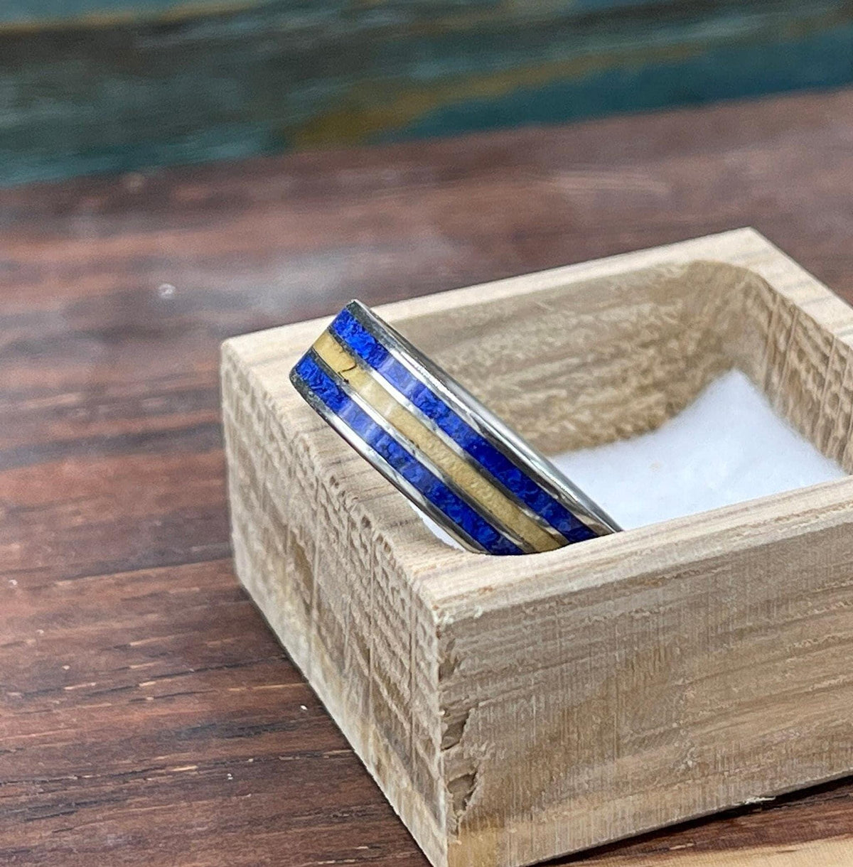 A titanium ring with a blue lapis lazuli and yellow wood inlay is displayed in a small, light-colored wooden box.