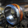 A close-up, angled view of a silver-colored ring with a wide, reddish-brown wood inlay, resting on dark mossy ground.