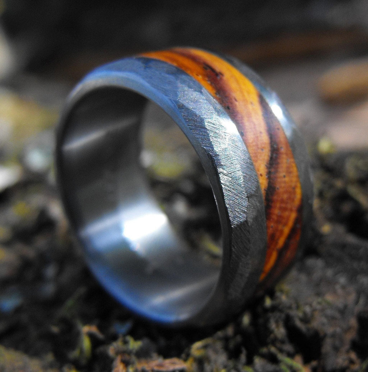 A close-up, angled view of a silver-colored ring with a wide, reddish-brown wood inlay, resting on dark mossy ground.