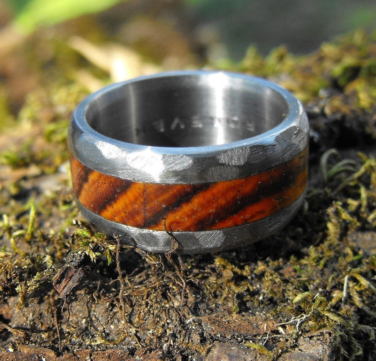 A close-up, straight-on view of a silver-colored ring with a wide, reddish-brown wood inlay, resting on dark mossy ground, with the word "FOREVER" engraved inside the band.