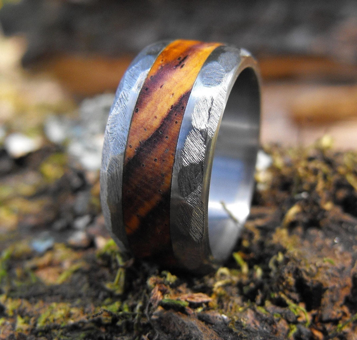 A close-up, angled view of a silver-colored ring with a wide, reddish-brown wood inlay, resting on dark mossy ground, with a bright highlight on the left side of the band.
