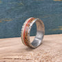 A silver-colored titanium ring with a textured band, featuring a central inlay of red opal flanked by light-colored wood, on a light wood surface.