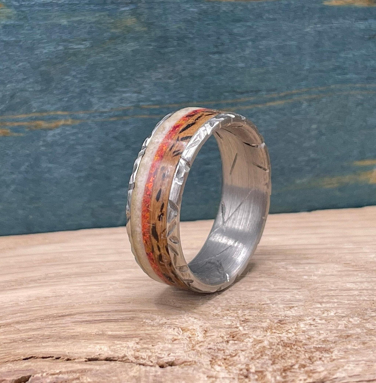A silver-colored titanium ring with a textured band, featuring a central inlay of red opal flanked by light-colored wood, on a light wood surface.
