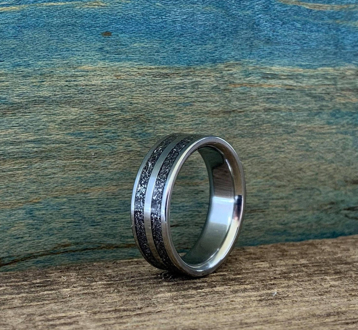 A silver-colored ring with two textured dark gray bands separated by a thin silver-colored stripe, on a wooden surface, viewed from a slightly different angle.