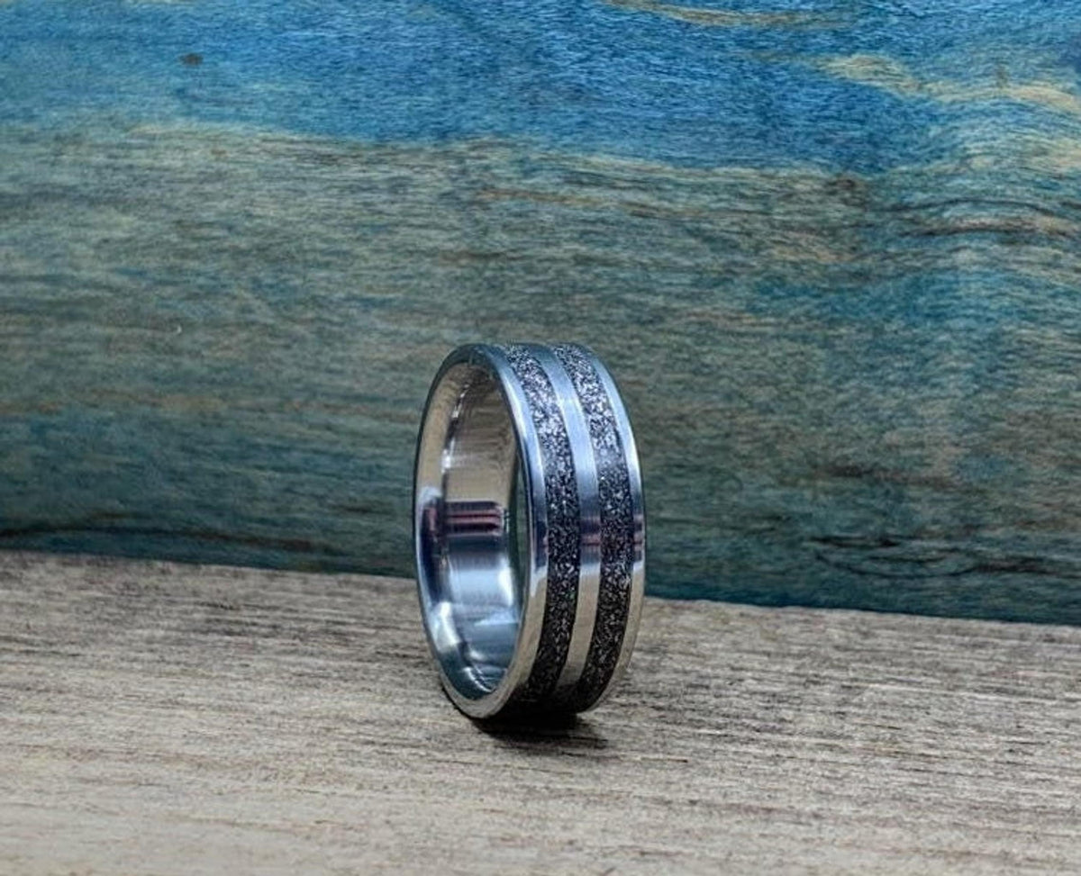 A silver-colored ring with two textured dark gray bands separated by a thin silver-colored stripe, on a wooden surface, viewed from a slightly different angle.