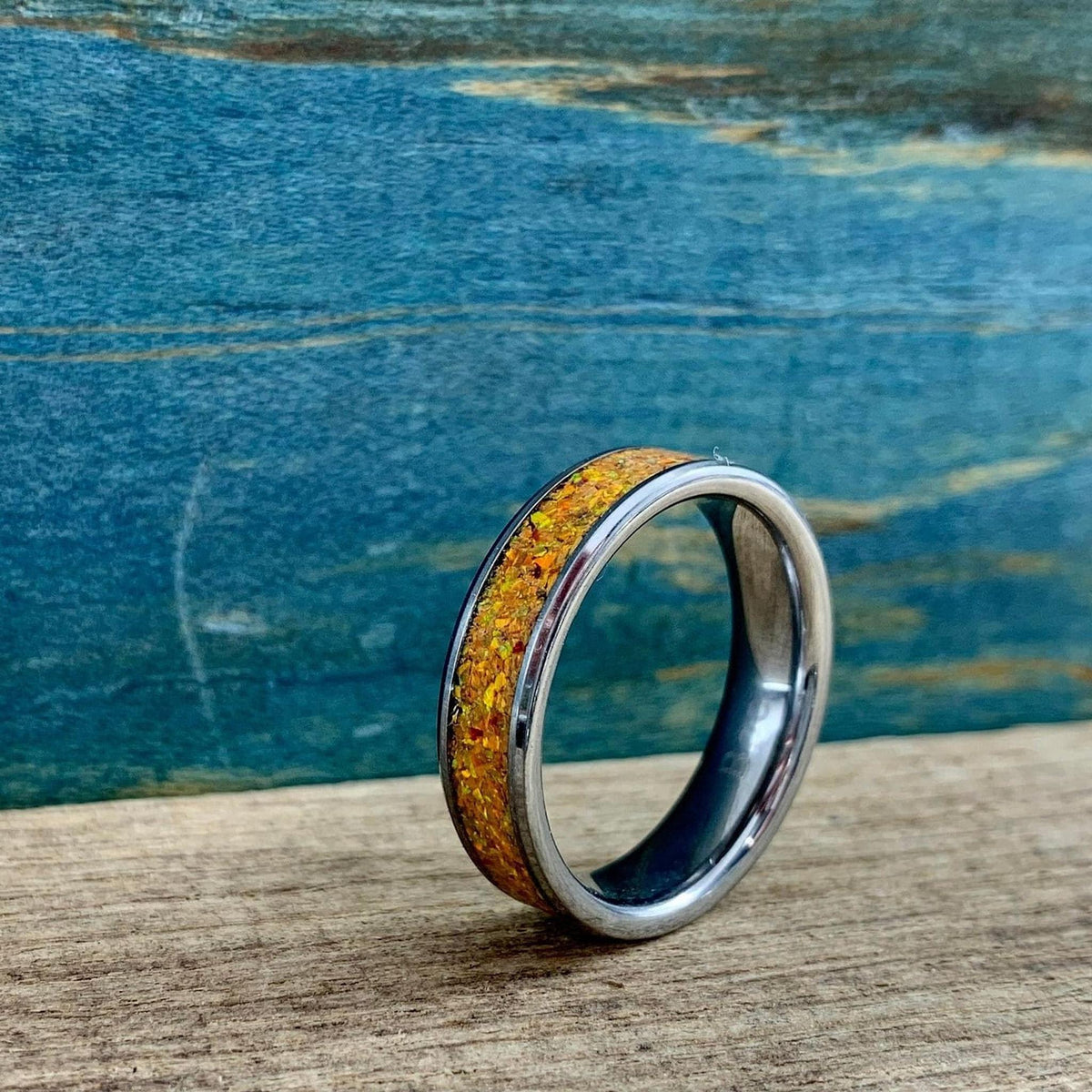 A silver-toned ring with an orange glittery inlay, viewed from the side on a wooden surface with a blue background.