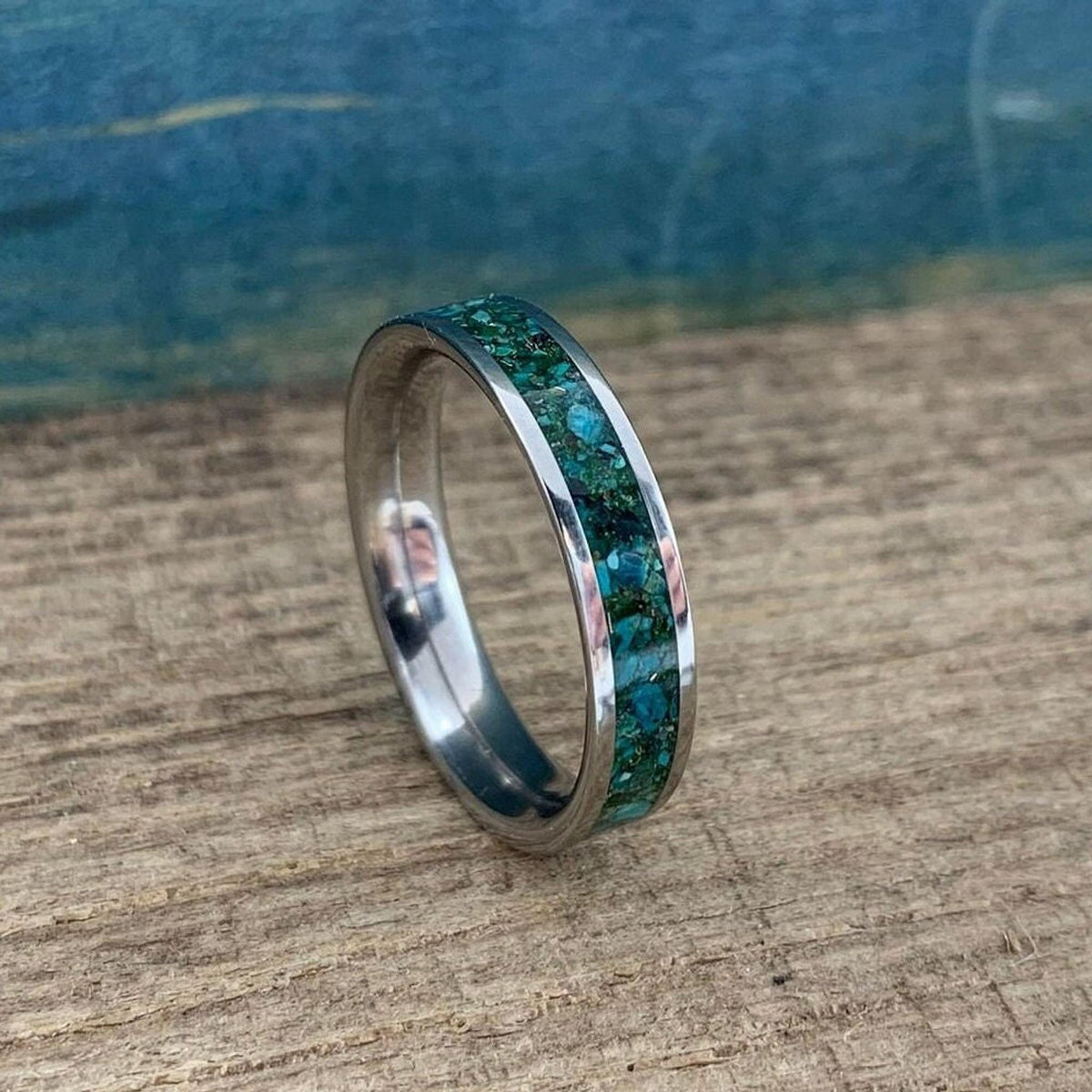 A silver-colored ring with a wide inlay of crushed turquoise and gold-colored flecks, shown at a slight angle on a wooden surface.