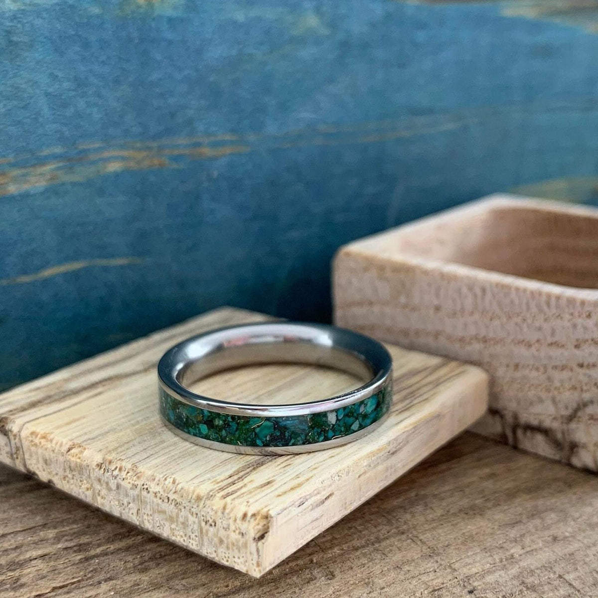 A silver-colored ring with a wide inlay of crushed turquoise and gold-colored flecks, resting on a light-colored wooden block on a wooden surface.