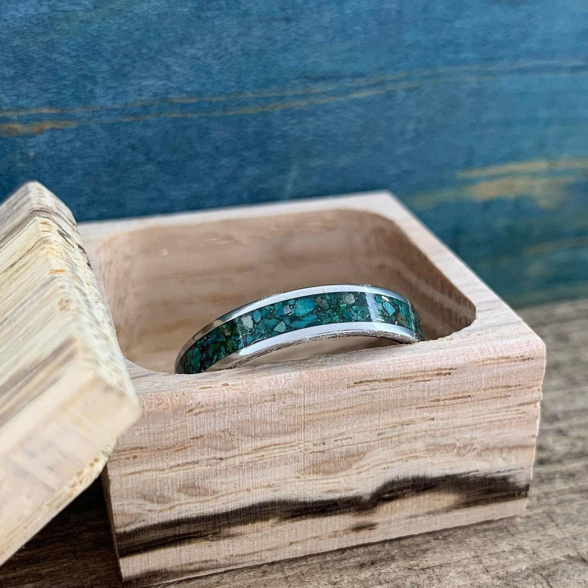 A silver-colored ring with a wide inlay of crushed turquoise and gold-colored flecks, resting inside an open light-colored wooden box.