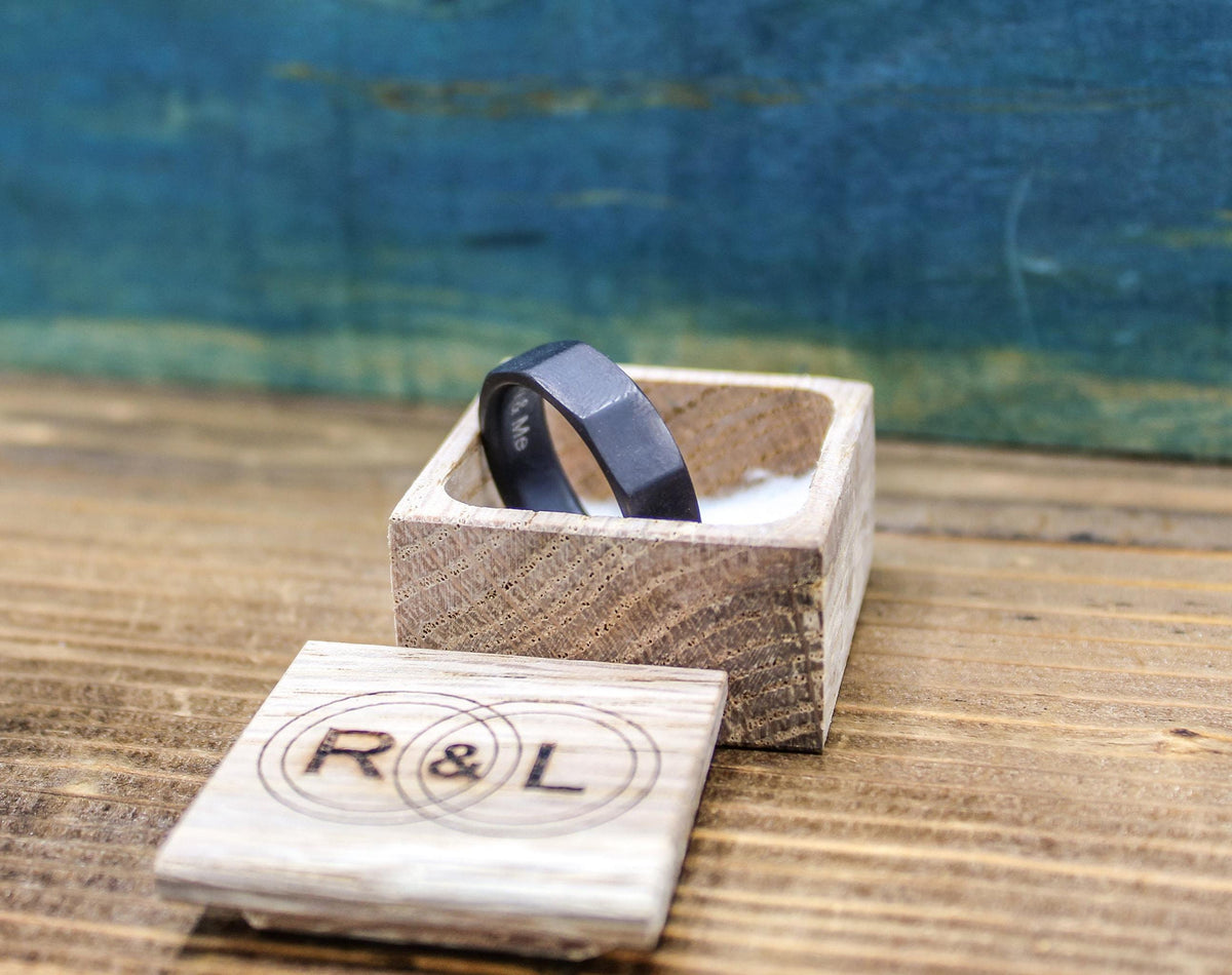 A dark gray, ten-sided ring with "R & L" and "To My Love" engraved on the inside rests in an open light wooden box, with a blurred blue background.