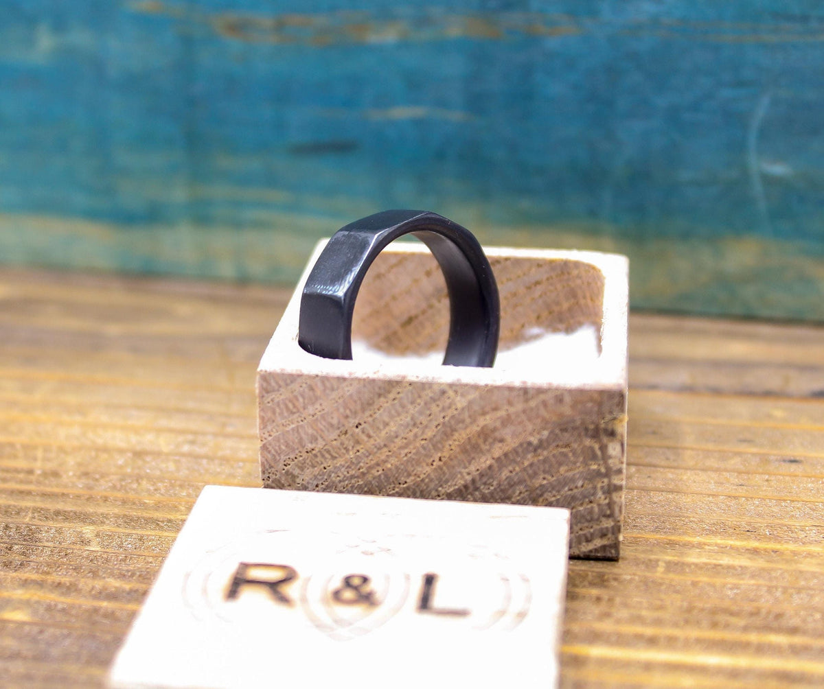 A dark gray, ten-sided ring rests inside an open light wooden box, with the lid showing "R & L" in front, and a blurred blue background.
