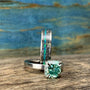 A silver-toned ring with a green square-cut stone sits in front of a wider silver-toned band with a black and blue-green inlay, all on a wooden surface.