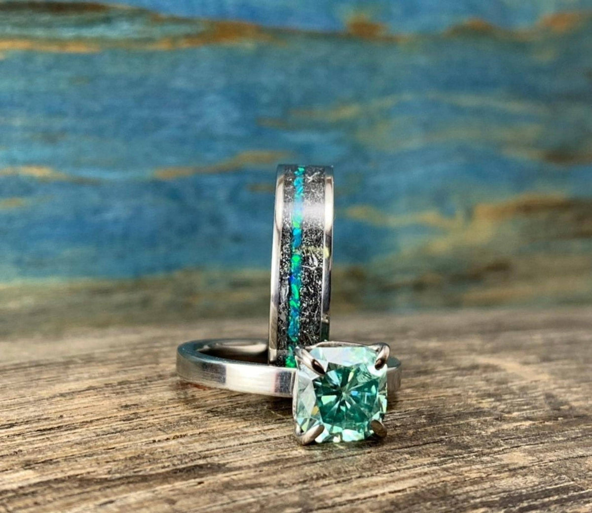 A silver-toned ring with a green square-cut stone sits in front of a wider silver-toned band with a black and blue-green inlay, all on a wooden surface.
