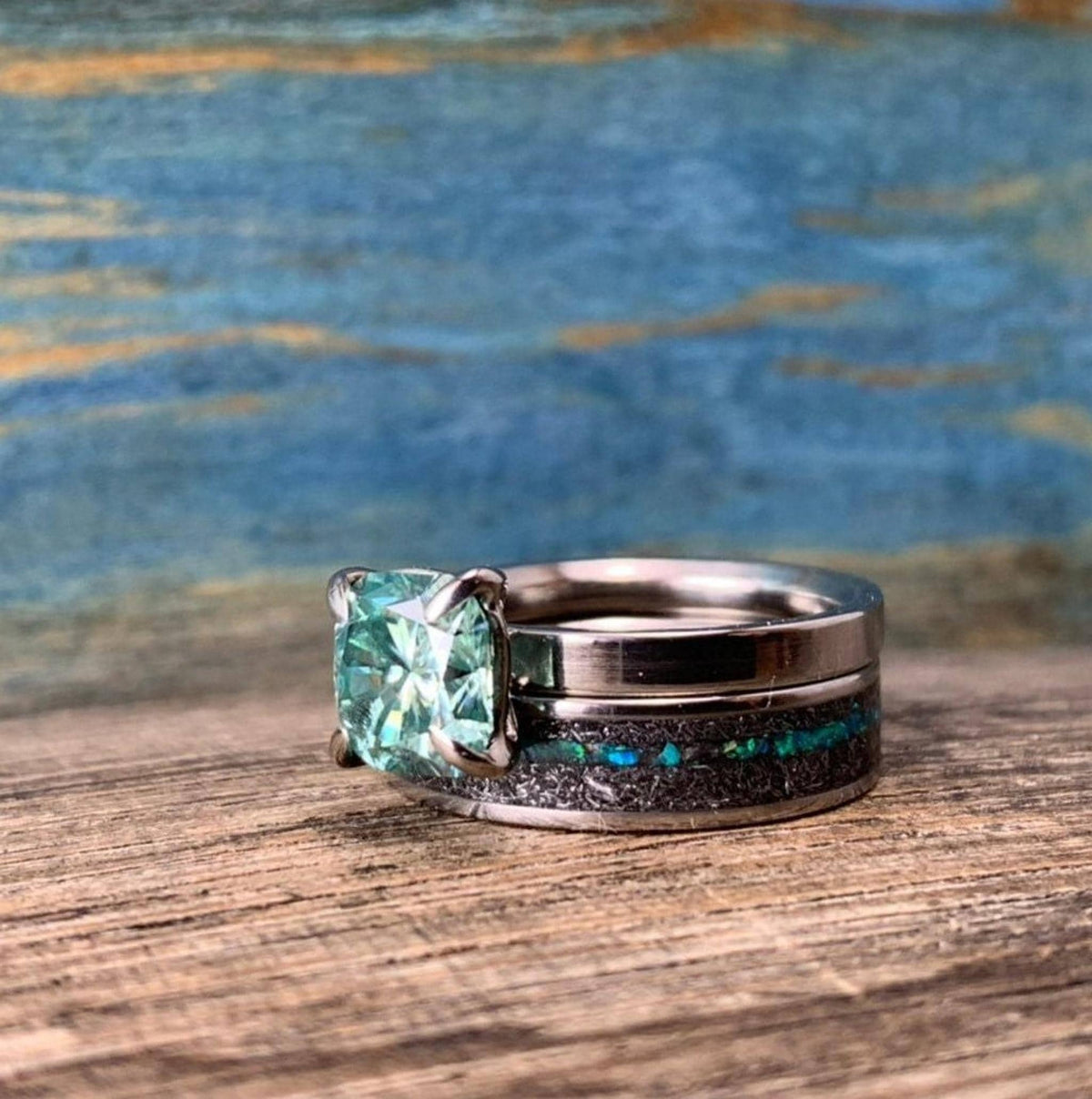 A silver-toned ring with a green square-cut stone is stacked on top of a wider silver-toned band with a black and blue-green inlay, on a wooden surface.