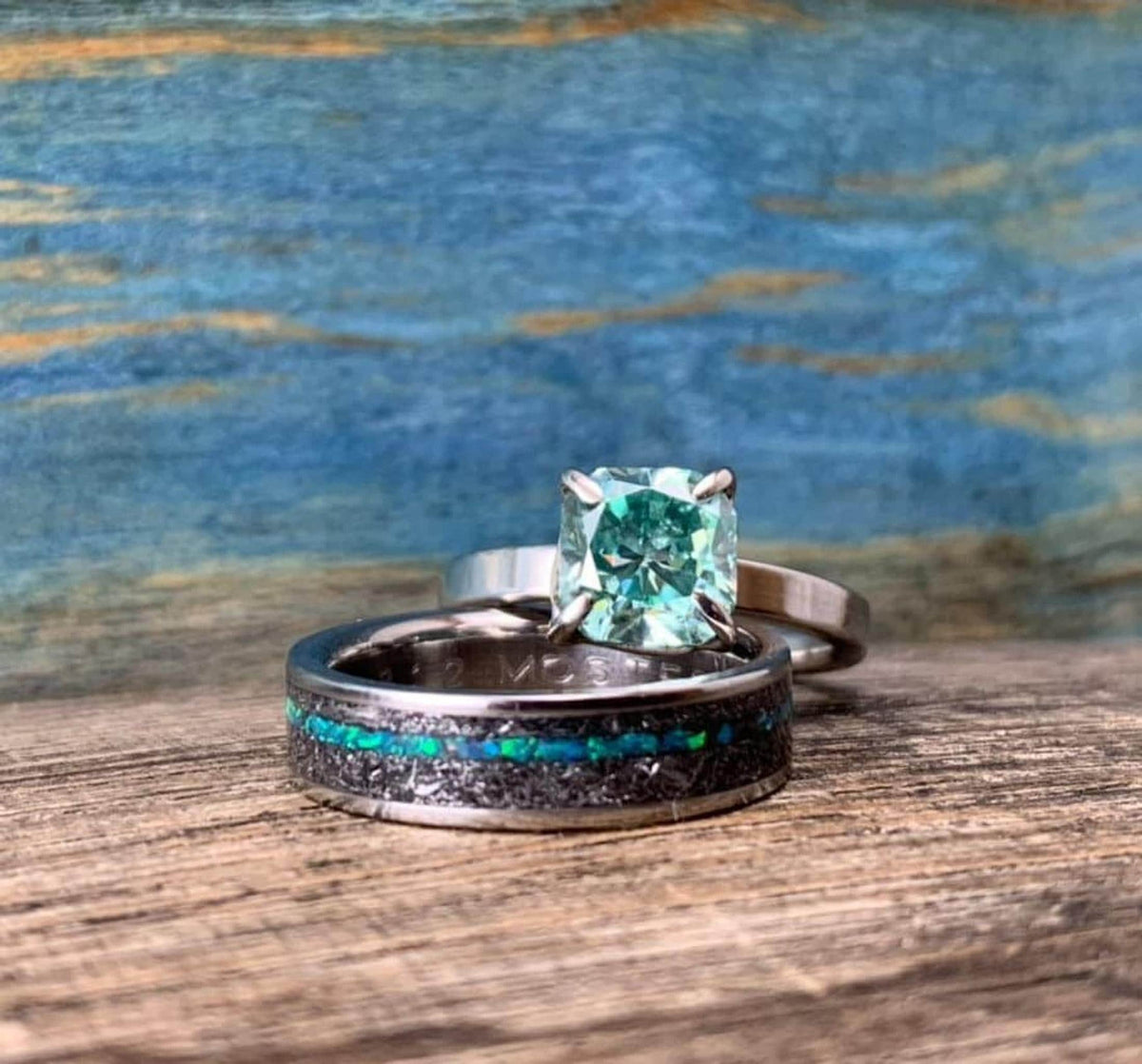 A silver-toned ring with a green square-cut stone is stacked on a wider silver-toned band with a black and blue-green inlay, on a wooden surface.